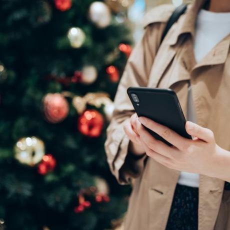 fotografie decupată și secțiunea mijlocie a unei femei care folosește un smartphone în fața unui brad de Crăciun colorat în sezonul festiv de Crăciun