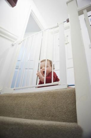 Poartă de siguranță pentru bebeluși pe scări: băiețel pe palierul casei sale. El se află în spatele unei porți de siguranță pentru copii care îl oprește de pericolul scării.