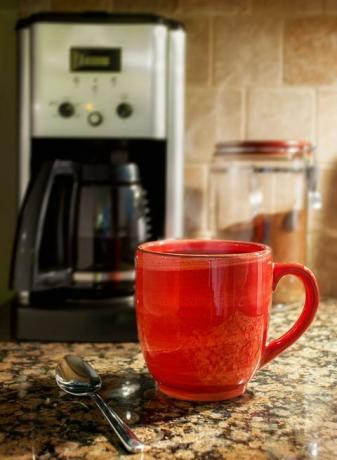 Ceașcă aburitoare de cafea: O cană roșie de cafea aburitoare se sprijină pe un blat de bucătărie din granit. Un filtru de cafea și un recipient de cafea măcinată sunt vizibile pe fundal.