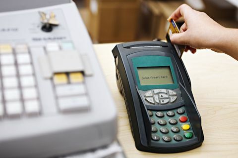 Mâna unei femei trece un card printr-o mașină de credit care stă lângă o casă de marcat. Concentrarea este pe mașina de credit.